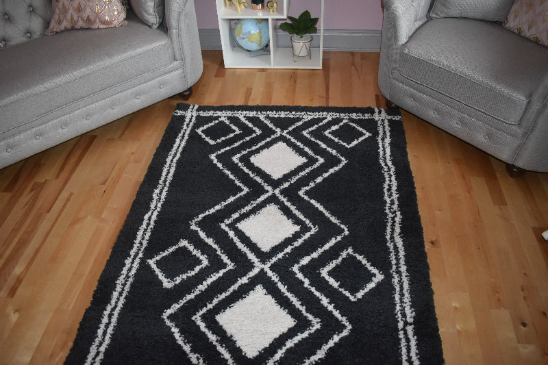 Black and white geometric patterned rug on a wooden floor with gray chairs and a small shelf in the background.