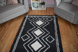 Black and white geometric patterned rug on a wooden floor with gray chairs and a small shelf in the background.