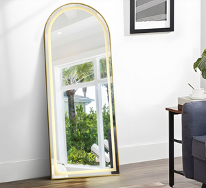 Full-length mirror with gold frame leaning against a white wall in a room.