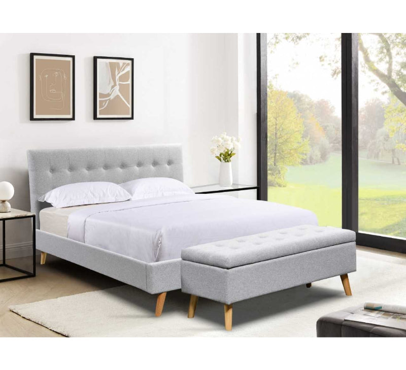 Grey upholstered bed with ottoman in a bright room with large windows.