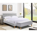 Grey upholstered bed with ottoman in a bright room with large windows.