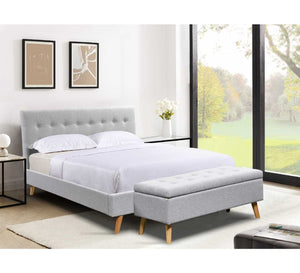 Grey upholstered bed with ottoman in a bright room with large windows.