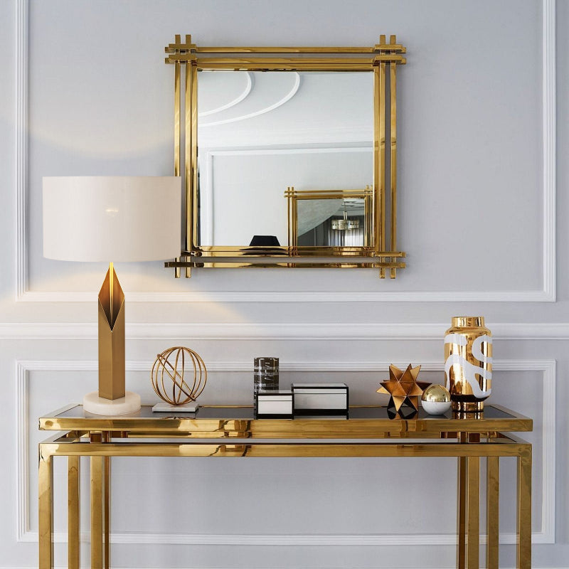 Decorative console table with gold mirror, lamp, and sculptures against a light gray wall.