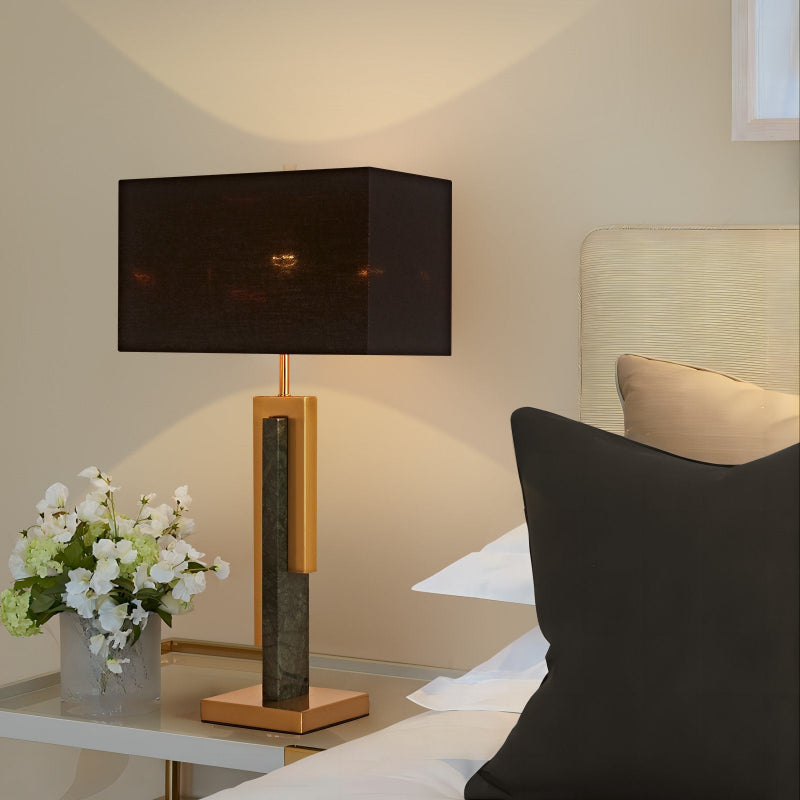 Decorative lamp with a black shade on a side table next to a bed with pillows.