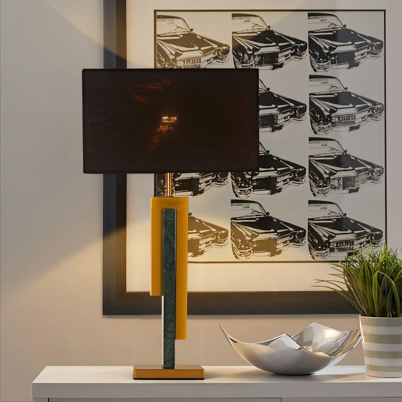 Table lamp with a black shade and yellow base on a table, with a framed artwork of cars in the background.