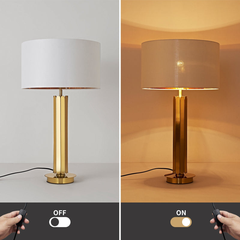 Table lamp with gold base and white shade on a gray background, switched on with warm glow.