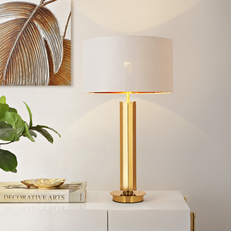 Gold table lamp with a white shade on a white surface, with a plant and decorative items in the background.