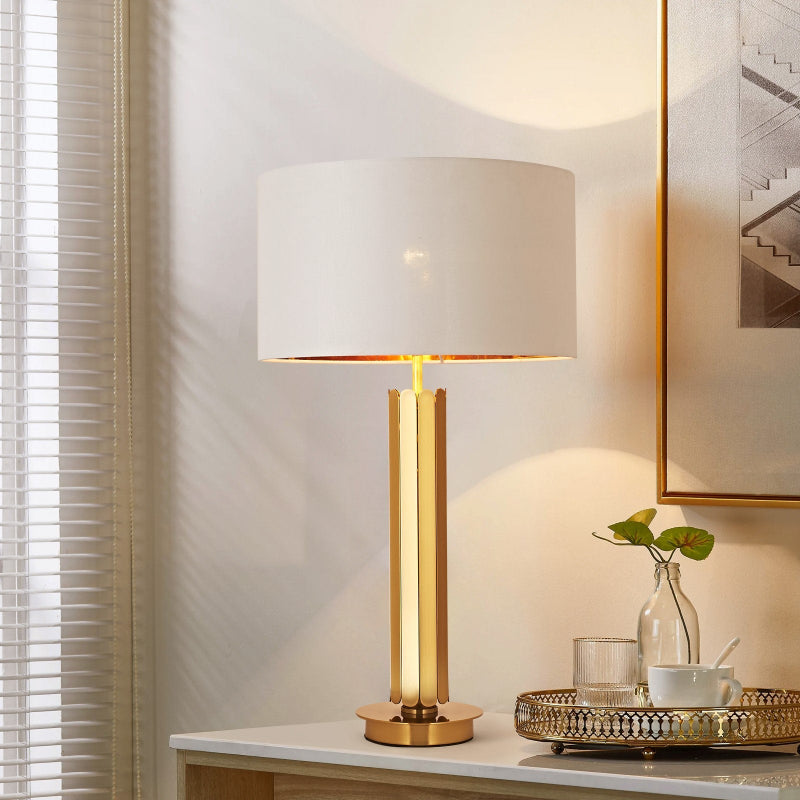 Decorative table lamp with a gold base and white shade on a surface near a window.