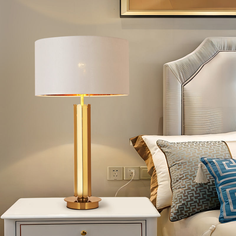 Modern table lamp with gold base and white shade on a nightstand next to a bed with decorative pillows.