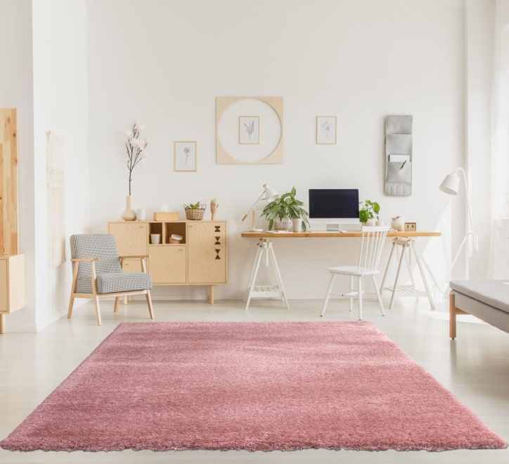 A plush pink shaggy rug on a white floor, with a beige wooden desk, chair, and armchairs in the background, suggesting a cozy living or office space.
