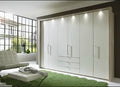 A modern white wardrobe with bi-fold doors and internal shelving, situated in a bright room with a chair and a small table in front.